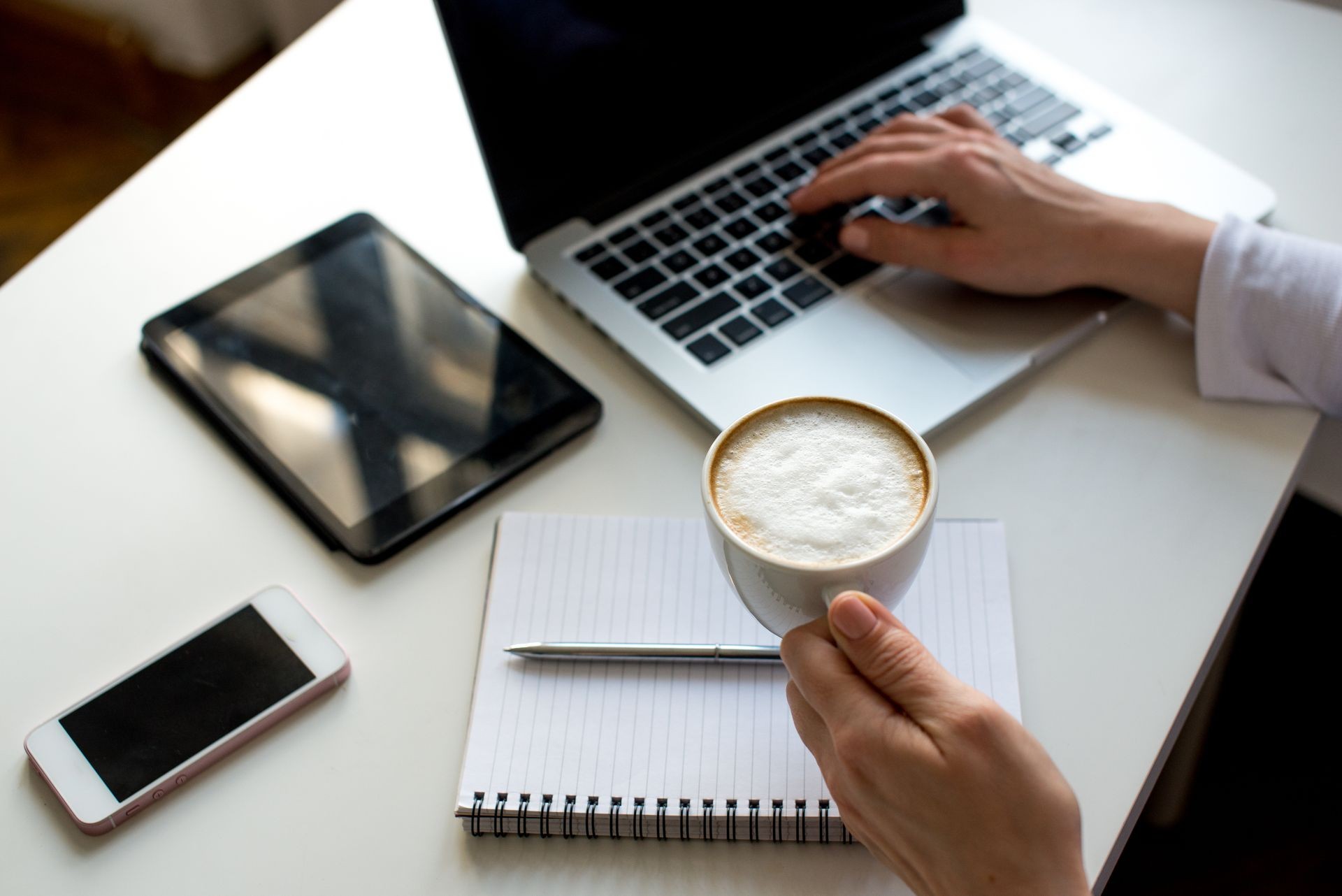 Young woman taking a coffee break as she sits at a table working on a laptop computer, tablet, phone, close up of her hands and the drink. Desing in white color. Buisness background